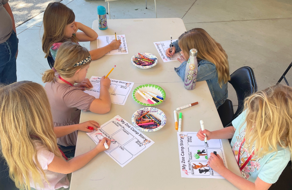 Children sitting around a table writing and coloring a "My Zoo Camp Story" worksheet.